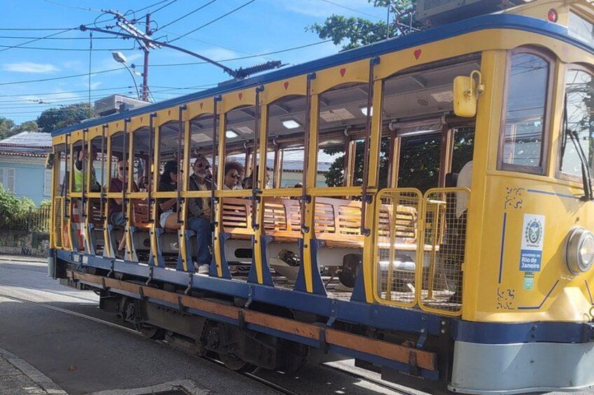 A yellow tram with passengers, parked on tracks, with overhead wires and buildings in the background. 3
