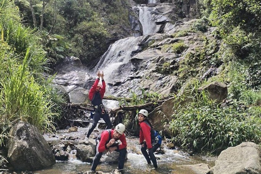 Three people crossing a shallow stream in a forested area with a waterfall in the background. 4