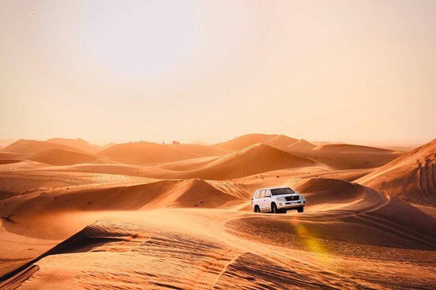 Un véhicule blanc traverse un paysage désertique, avec des dunes de sable ondulantes à l’horizon. 1