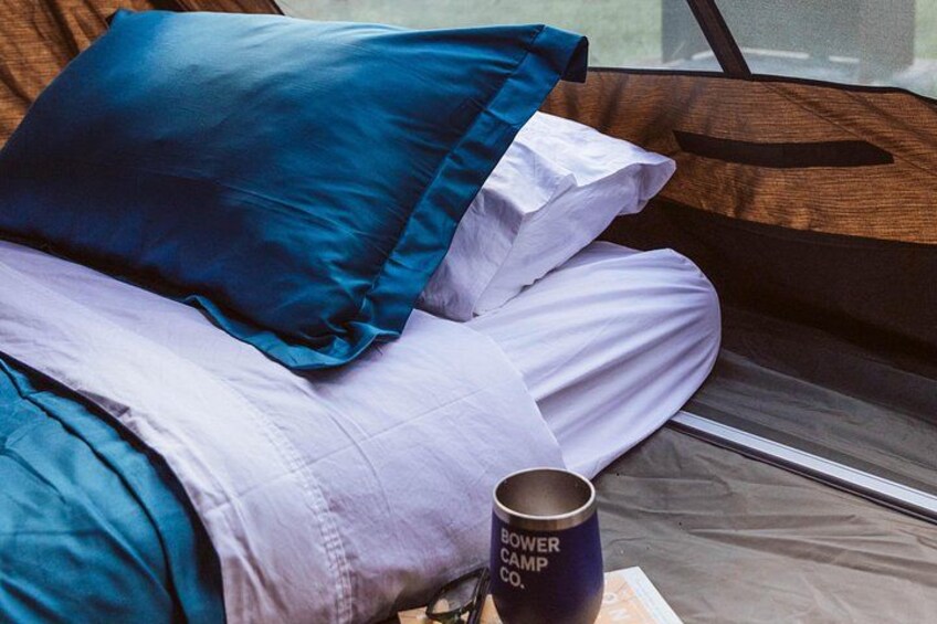 Una cama con almohadas azules y blancas, una taza con el texto 'BOWER CAMP CO.' y una ventana. 3