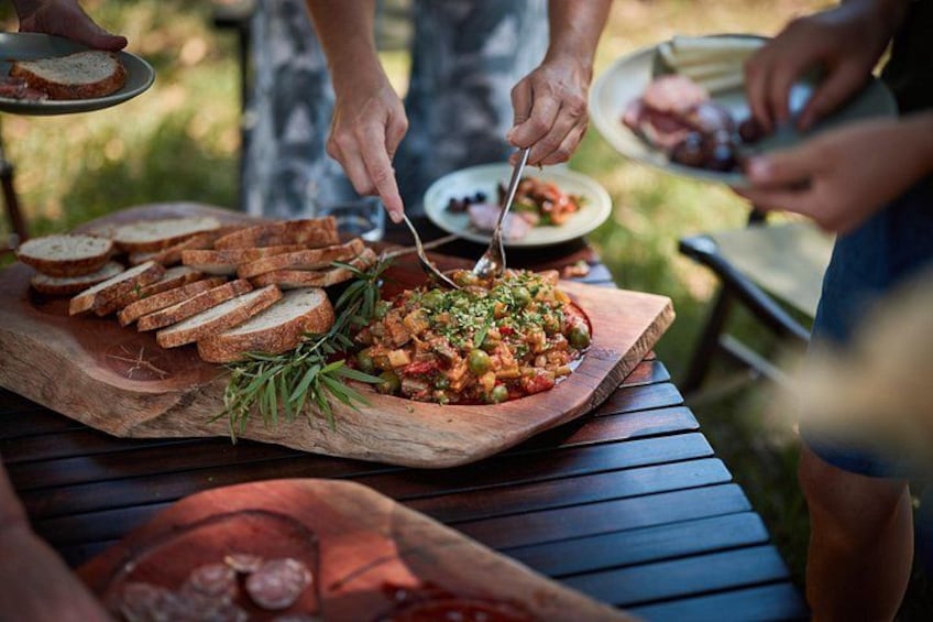Una tabla de madera con pan rebanado y una ensalada, un plato con carne, y una persona sirviendo comida. 4