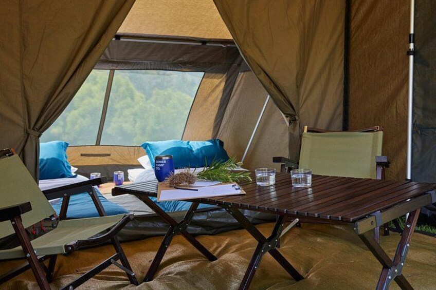 Una carpa con una mesa y sillas en su interior, una zona de descanso con cama y almohadas, y vistas a árboles desde afuera. 1