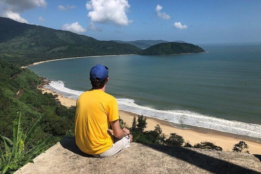 A man in a yellow shirt and blue cap sitting on a ledge overlooking a beach with waves, greenery, and distant hills. 2