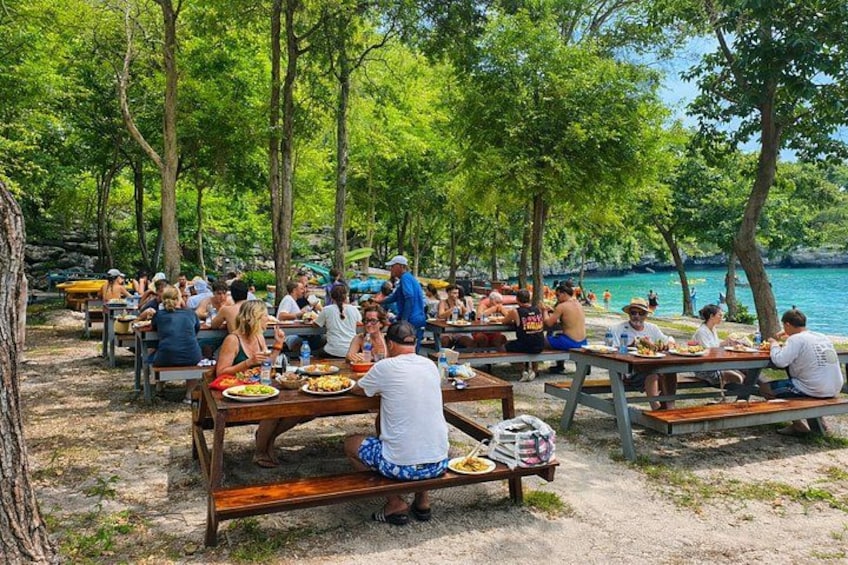 People dining outdoors at a lakeside picnic area with tables and benches. 1