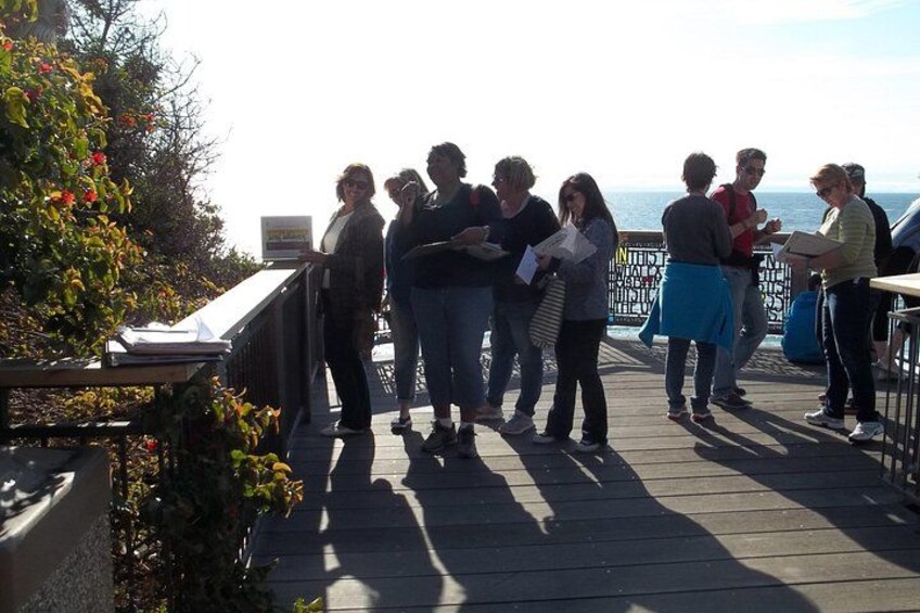 En gruppe mennesker står på en træterrasse ved havet og taler sammen. 4