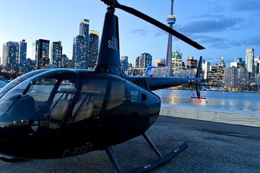 En helikopter med registreringsnummer G-FKX står på en helikopterplatta, med stadens silhuett och CN-tornet i bakgrunden. 1