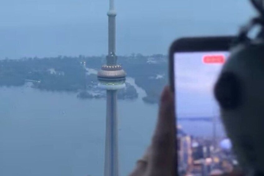A person taking a photo of a tower with a circular observation deck, surrounded by water and distant land. 1
