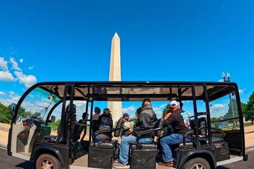 A black vehicle with passengers, a clear blue sky, and a tall monument in the background. 2