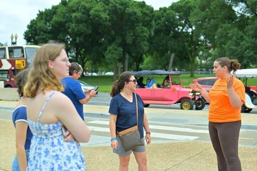 A group of people standing on a sidewalk, a red car, and a green park in the background. 2