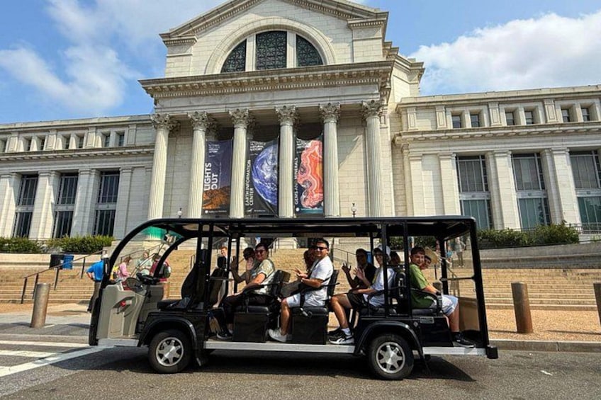 Een zwarte touringcar met passagiers, geparkeerd voor een groot klassiek gebouw met kolommen en een koepel. 3