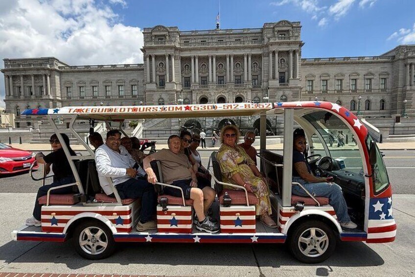 A small, open-top vehicle with American flag decorations, parked on a street with a large building in the background. 1
