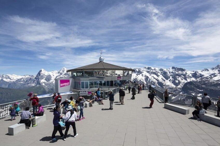 A mountain observation deck with people walking and sitting, overlooking snow-capped peaks. 1