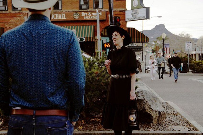 A person in a blue patterned shirt and a hat, a person in a black dress with a lantern, and a street with buildings and a mountain in the background. 4