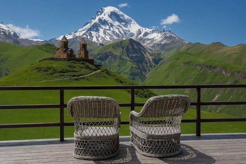 Korbstühle auf einer Holzterrasse mit Blick auf eine gebirgige Landschaft und ein historisches Gebäude. 1