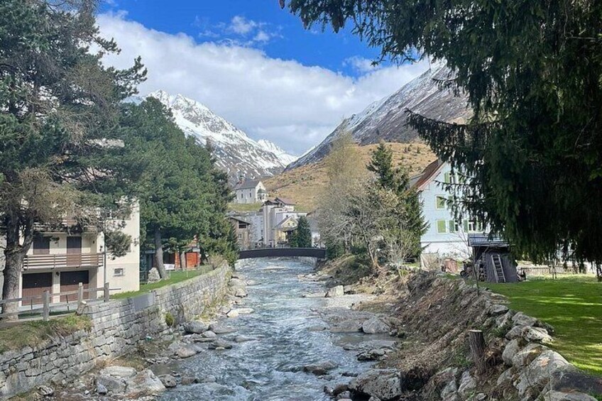 A river flowing through a valley with houses on either side, a bridge in the distance, and snow-capped mountains in the background. 4