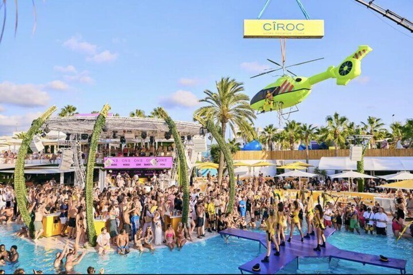 A crowded outdoor event with a pool, palm trees, and a helicopter. 3
