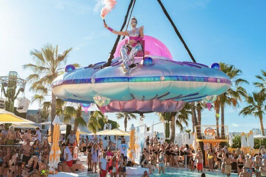 A person performing on a large, colorful UFO-like float above a crowded pool. 5