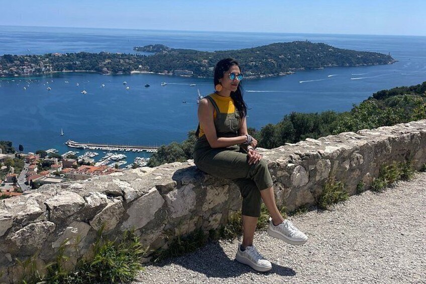 A person sitting on a stone wall overlooking a coastal town with boats and a distant island. 4