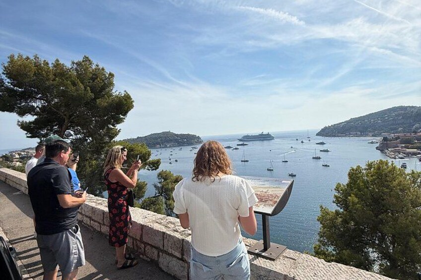 A coastal view with people standing on a stone wall, overlooking a bay with boats and a distant island. 3