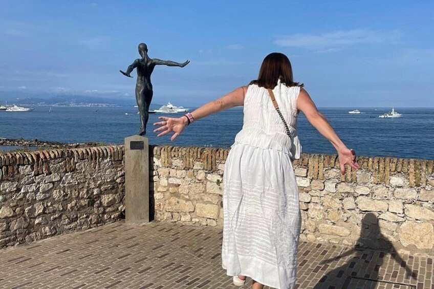 A woman in a white dress standing on a stone wall overlooking the sea with a statue and boats in the background. 2