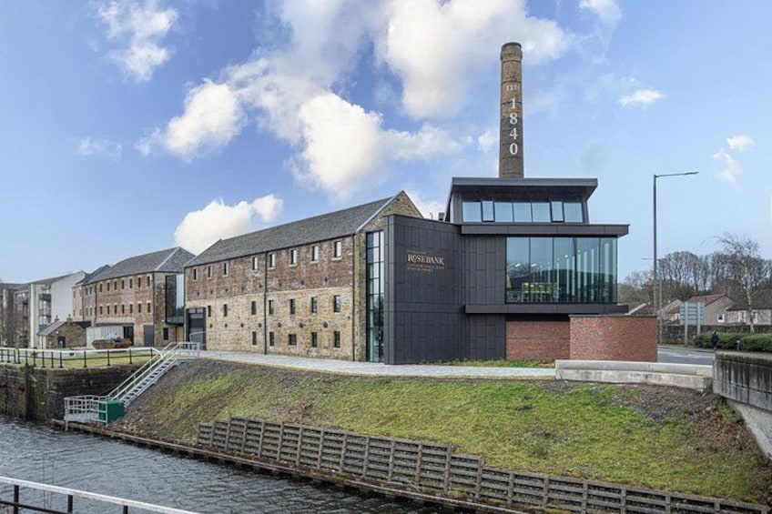 A historic building with a tall chimney, a modern extension, and a river in the foreground. 2
