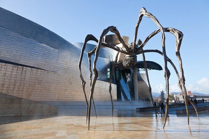 A modern building with a reflective surface and a large metallic sculpture resembling a spider. 1