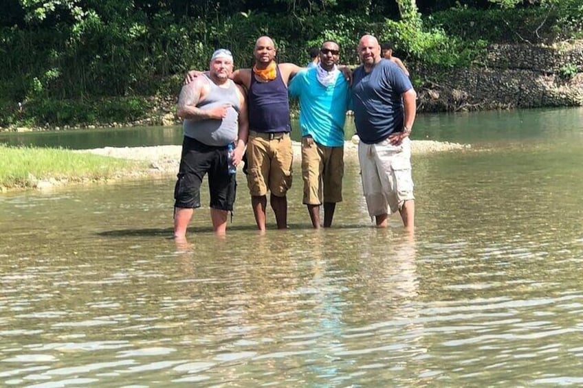 Een groep mensen staat in ondiep water langs een rivieroever, met begroeiing op de achtergrond. 3