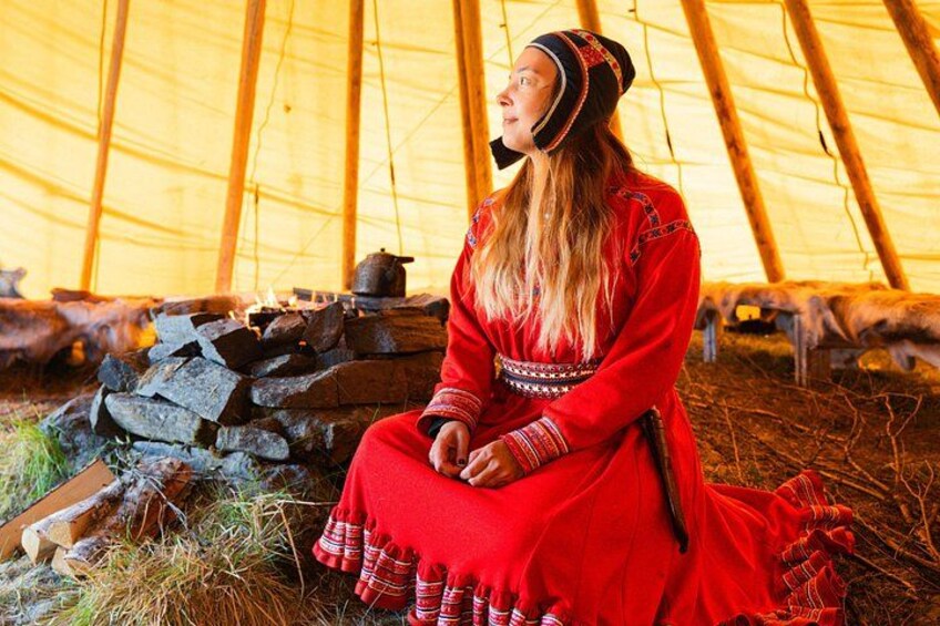 Una persona vestida con atuendo rojo tradicional sentada dentro de una tienda de campaña, con un fuego y leña cerca. 3