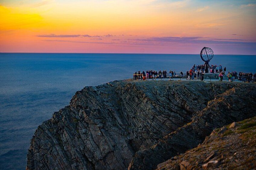Costera rocosa con una estructura esférica en la cima, un atardecer de fondo y una multitud de personas reunidas al borde. 1