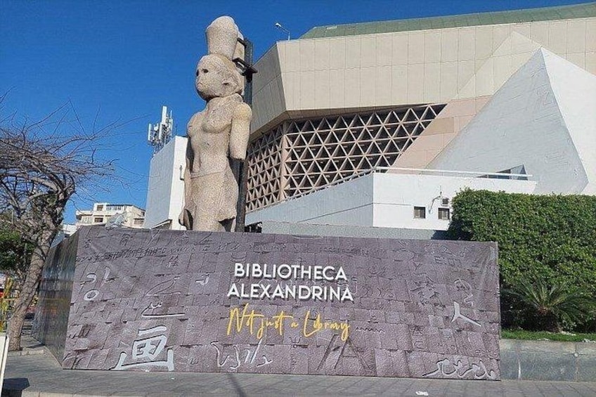 A modern building with a unique geometric design, a large stone sculpture, and a sign for the Biblioteca Alexandrina. 1