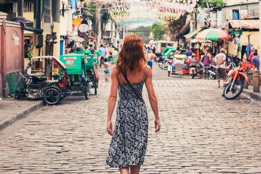 A woman in a patterned dress walking down a cobblestone street with shops and vehicles. 1