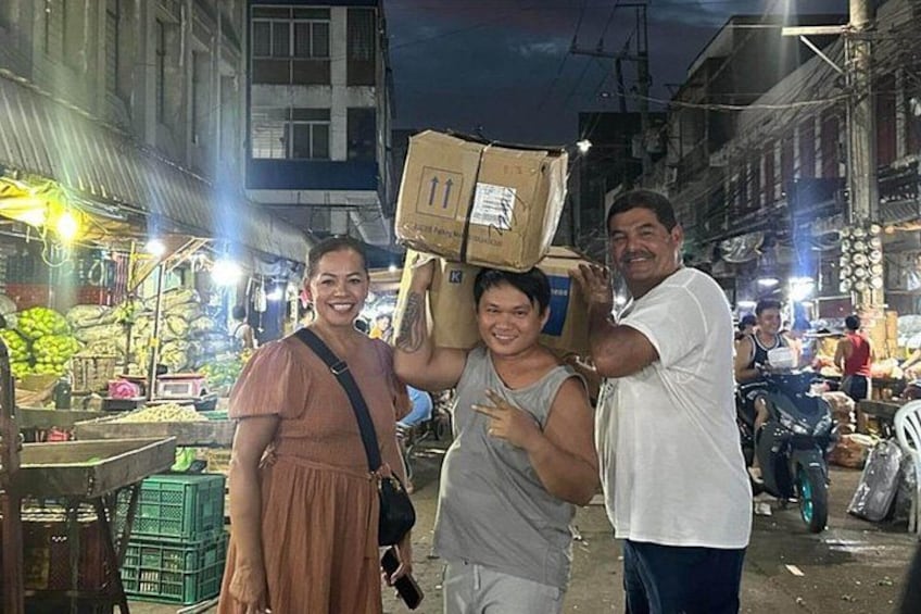A nighttime street scene with people carrying boxes, market stalls, and buildings in the background. 2