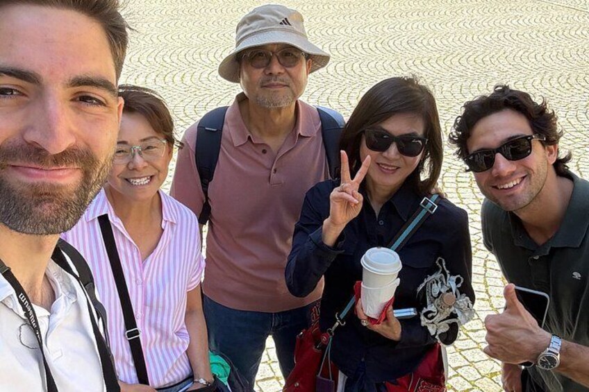 A group of people outdoors, some holding a coffee cup and a camera, with a patterned ground in the background. 2