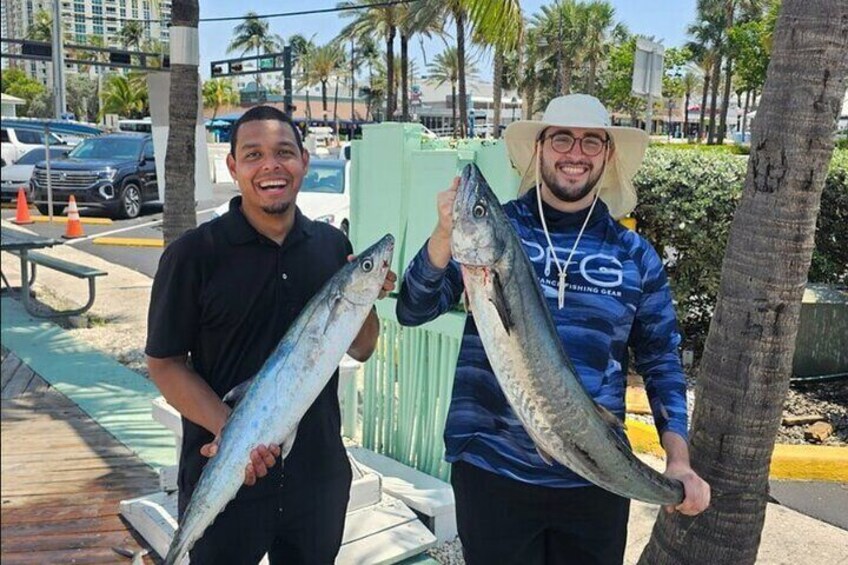 Two people holding large fish, a green fence, and palm trees in the background. 5