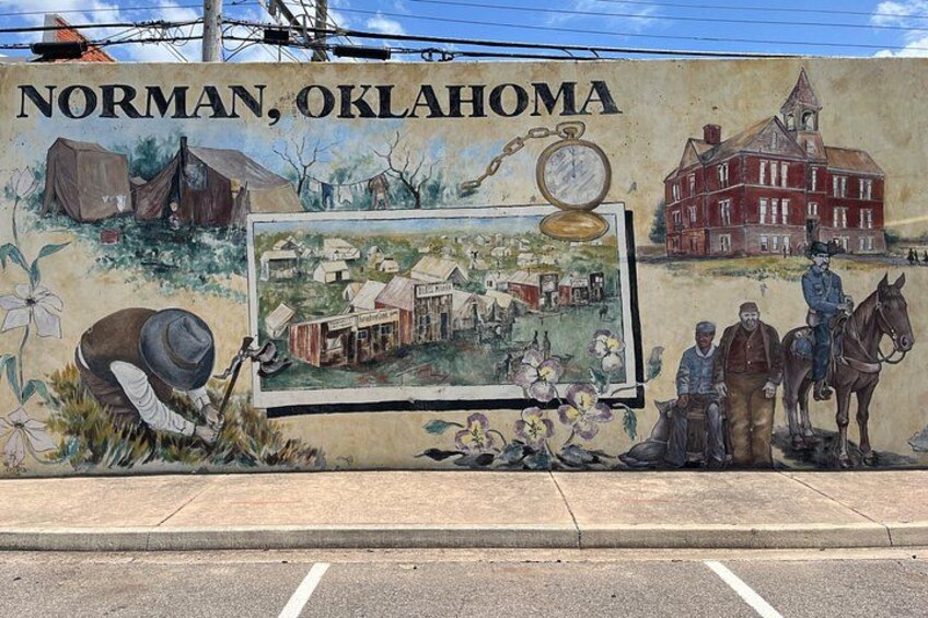 A mural on a wall depicting Norman, Oklahoma, with historical buildings, a horse, and a man with a pocket watch. 1