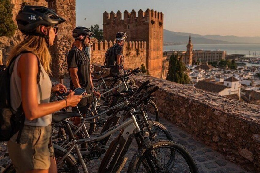 Un grupo de ciclistas cerca de un muro de piedra histórico con vistas a un paisaje urbano y un castillo al fondo. 3
