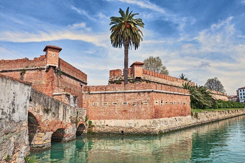 A historic brick fortification with a palm tree in the foreground, a bridge, and a calm water body. 2