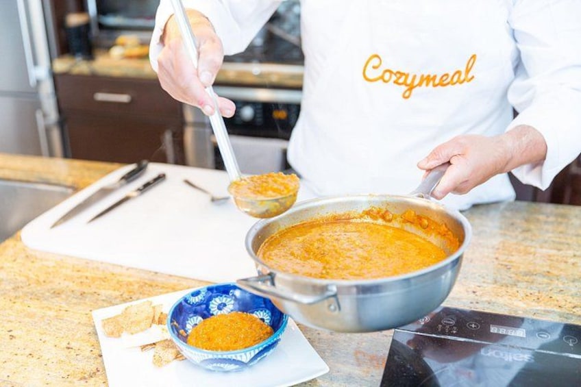 A person in a white chef's coat with 'Cozymeal' text, stirring a pot of orange-colored food on a kitchen counter. 3