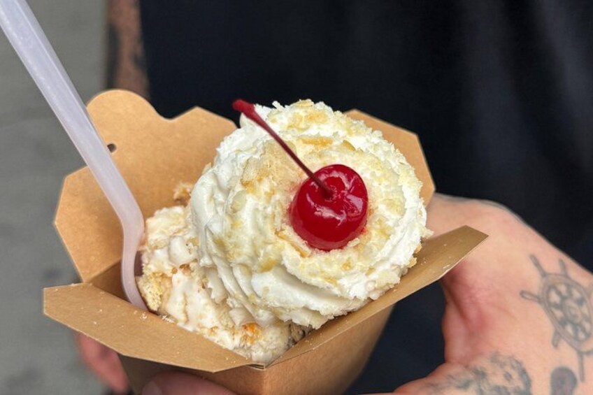 Una persona sosteniendo un recipiente de cartón con un postre cubierto de crema batida y una cereza. 3