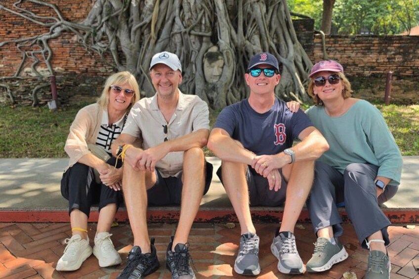 Four people sitting on a bench with a large tree and brick wall in the background. 4