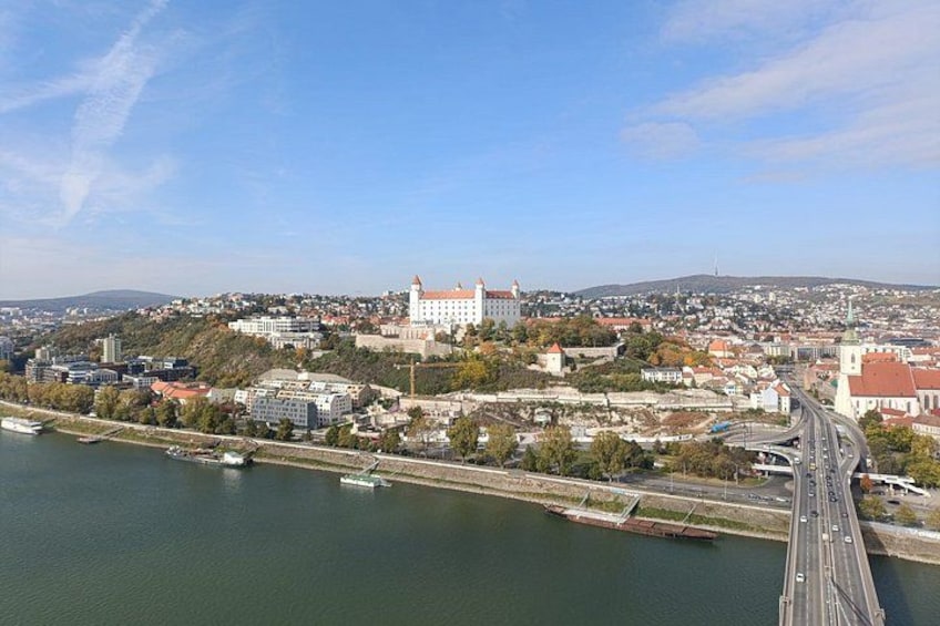 A river with a bridge, a large white building with a red roof, and a hilly landscape in the background. 1