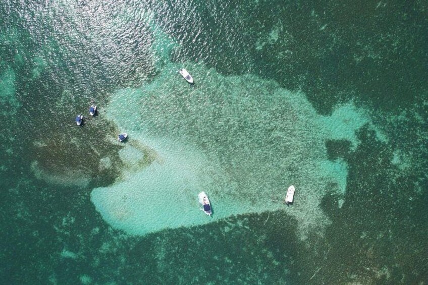 An aerial view of a coastal area with clear turquoise waters, several boats, and a small island with dense vegetation. 1