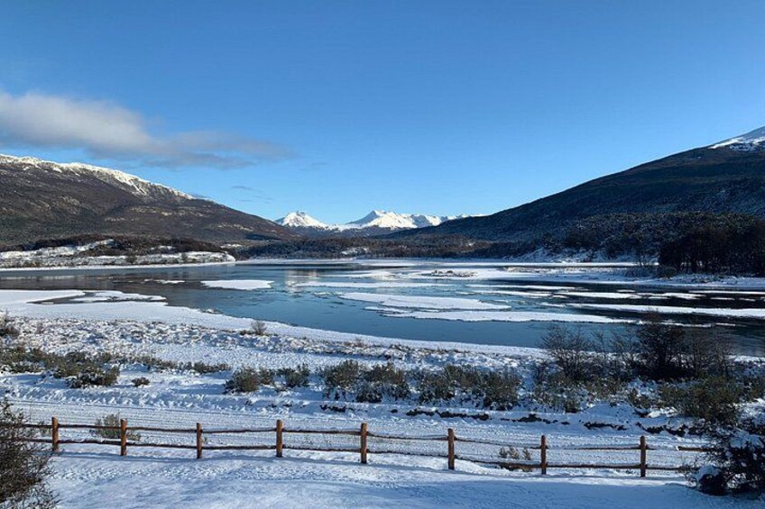 A frozen river with snow-covered banks, a wooden fence, and mountains in the background. 2