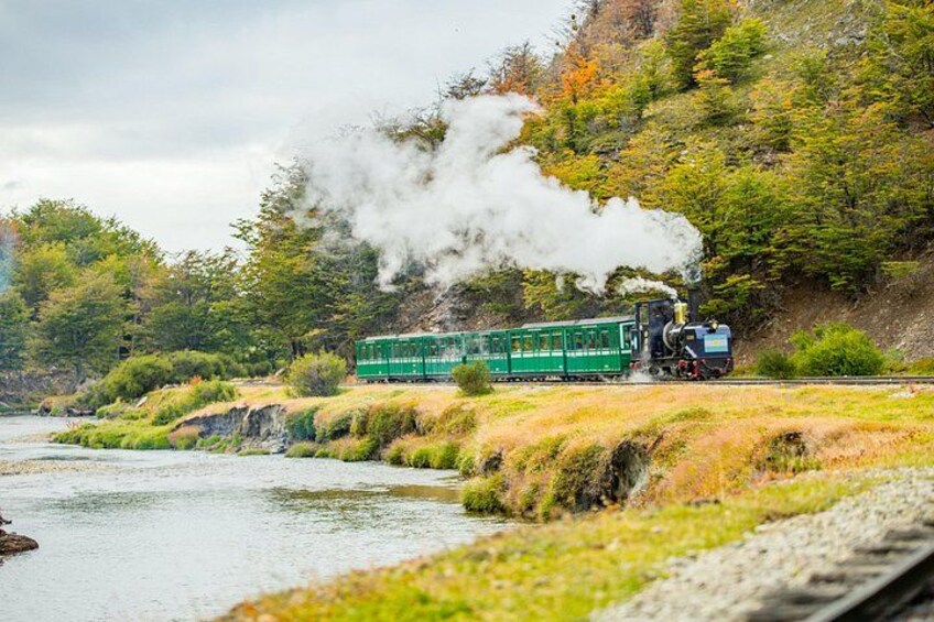 Un treno verde con vapore che esce dal motore, che viaggia lungo un binario accanto a un fiume, circondato da alberi dai colori autunnali. 5