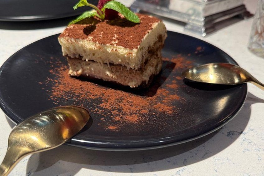 En skive tiramisu på en mørk tallerken med en ske og et drys kakaopulver. 2