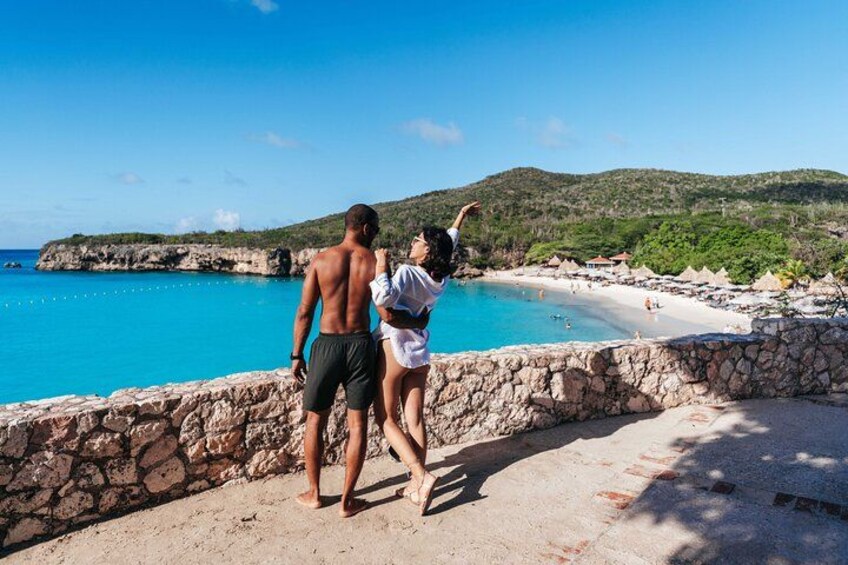 A couple standing on a stone wall overlooking a beach with clear blue water and a hilly landscape in the background. 4