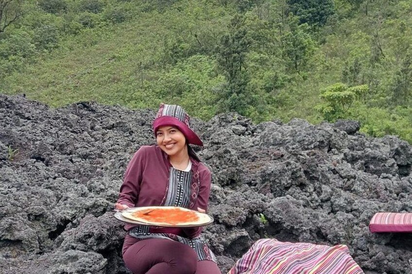 Une personne assise sur une surface rocheuse, avec une assiette de nourriture devant elle, entourée de roches volcaniques et de végétation. 2
