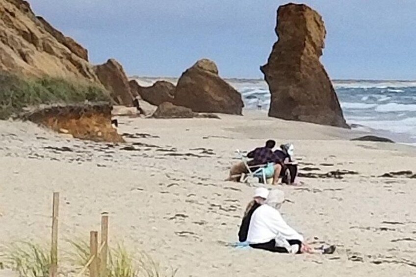 A sandy beach with large rock formations and people sitting on the sand. 2