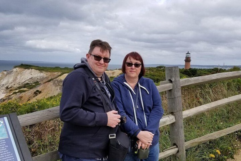 Two people standing by a wooden fence with a coastal landscape and a lighthouse in the background. 4