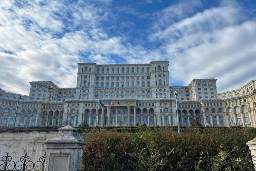 Ein großes, historisches Gebäude mit mehreren Stockwerken und Rundbogenfenstern, umgeben von Grün und unter einem teils bewölkten Himmel. 1
